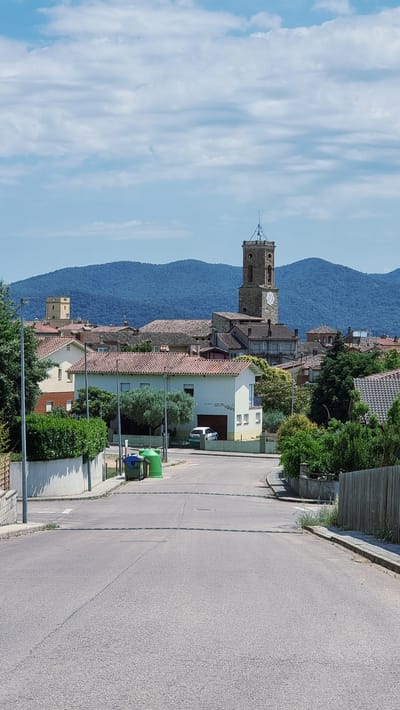 The image depicts a sunlit, upward-angled view of a village street leading towards a cluster of buildings in Montagut i Oix, Spain. Dominating the background are rolling, tree-covered blue hills under a sky filled with white, wispy clouds. A prominent stone bell tower with a clock face rises above the red-tiled roofs of the houses in the middle ground. In the foreground, the asphalt street curves gently, lined with green hedges and a sidewalk. Two green recycling bins are situated on the sidewalk. A white car is parked in front of a house with a white facade and a garage door. The environment appears to be a peaceful residential area. There are no people visible in the image.