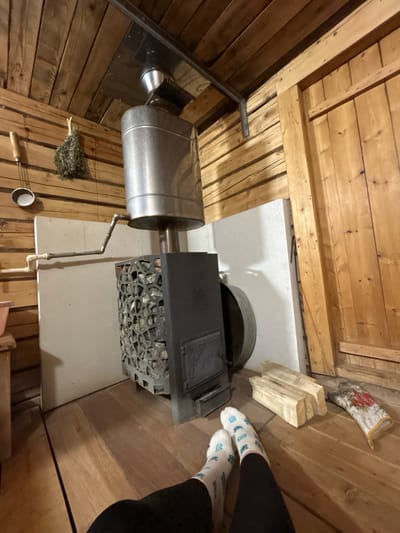 An interior shot shows a sauna stove with a cylindrical metal water tank connected by pipes, surrounded by wood paneling. A metal scoop, birch branches, and a glove sit near the stove. Wood logs are stacked beside the stove. Two people in socks and black pants stand near the stove. The city of Bashur, Russia is not discernible from the image.