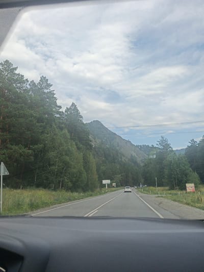 A frontal view from inside a vehicle shows a two-lane asphalt road traveling through a mountainous, forested area. The sky is overcast with scattered clouds. On the left side of the road, a dense forest of pine and deciduous trees lines the base of a steep, rocky mountain. On the right side of the road, there is a grassy verge with scattered trees and a few signs. A white car is visible in the distance, traveling in the same direction as the viewer. The road appears to be in a rural or semi-rural area, possibly on the outskirts of Uznezya, Russia. The interior of the vehicle is visible in the foreground, with a dark gray dashboard.