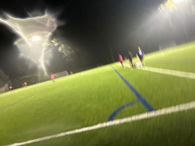 The image depicts an artificial turf sports field at night in Newton, United States. The scene is illuminated by multiple stadium lights, creating significant lens flare, particularly in the upper left quadrant. The field features bright green turf with distinct white and blue markings, including a prominent blue line extending diagonally from the lower center. A white goalpost structure is partially visible in the mid-ground on the left. Approximately six to seven blurry human figures are present on the field. Three figures are grouped on the left side, each wearing a pink shirt. Three additional figures are located closer to the right side: one in a blue/purple top, one in a dark top, and one in a pink top. The individuals appear to be standing on the field, with no specific actions or interactions discernible due to the image's blur and low resolution.