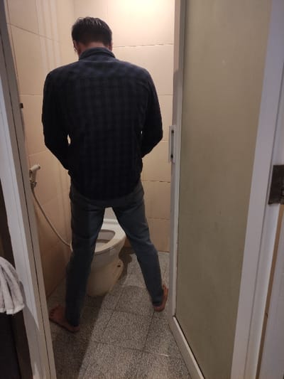 The image depicts a man in a bathroom, standing on a tiled floor. He is wearing blue jeans and a dark plaid shirt. The man is using a white toilet. The man is standing with his back to the camera, partially obscured by a closed white door, with a small portion of a white towel visible on the floor below. The bathroom is lit by natural light and the scene appears to be in a residential dwelling.
The coordinates provided -8.434, 114.18 - are located in the city of Gambiran, Indonesia.