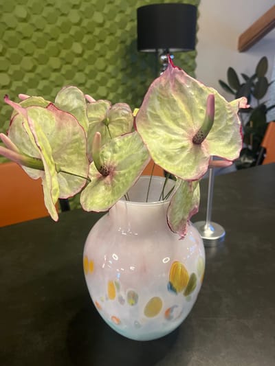 In a modern interior setting, possibly in Achadara, Abkhazia, a clear glass vase with a pale pink and white iridescent finish, decorated with multi-colored circular splotches, holds a bouquet of light green and pink-tinged anthurium flowers. The flowers, with their distinctive spathes and spadices, appear slightly wilted, their edges tinged with a darker pink or red hue. They are arranged to spill over the rim of the vase, creating a dynamic visual. The vase rests on a dark, reflective surface, likely a table. In the background, a textured green wall, resembling hexagonal tiles, occupies the upper left portion of the frame. To the right, a black lampshade with a metallic base is visible, and further in the background, a blurry silhouette of a plant and a wooden beam can be discerned. The lighting suggests an indoor environment with ambient light.
