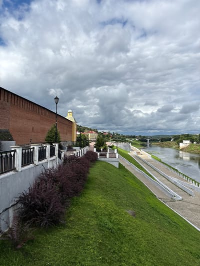 A scenic view of the embankment in Demenshchina, Russia, features a prominent red brick wall on the left, likely part of a historical fortification, with a black wrought-iron fence along its top. A paved walkway runs parallel to the wall, bordered by a grassy slope. Further to the right, a wide river flows, with a modern bridge visible in the distance. The embankment on the opposite side of the river is lined with buildings and lush green trees. The sky is filled with dramatic, dark clouds, suggesting an overcast day. No people are visible in the scene.