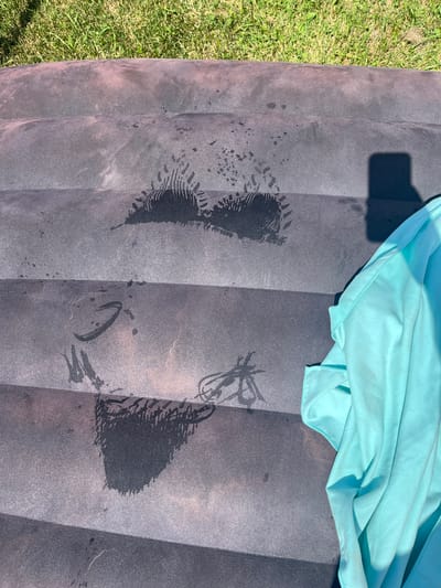 The image shows a close-up, overhead view of a dark, textured surface with several damp impressions. The surface appears to be made of a synthetic material, possibly an inflatable mattress or lounge chair, with horizontal ridges. Green grass is visible in the background. In the foreground on the right, a crumpled light blue towel or garment is partially visible, casting a shadow. The damp marks on the dark surface resemble the impressions left by fabric, possibly a bathing suit or towel, with some showing intricate patterns that suggest lace or textured material. There are also smaller water droplets scattered across the surface. The lighting suggests it is a sunny day. The location is identified as the city of Pushkino, Russia, though no specific urban features are visible to confirm this.