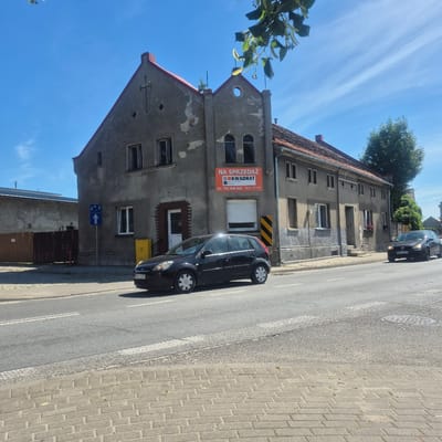 The image shows a street scene in Bralin, Poland, on a bright, sunny day with a clear blue sky and wispy clouds. In the foreground, a black car with license plate "PKE CS75" is parked on the right side of the road. The car is a hatchback, possibly a Ford Fiesta. The road surface is asphalt, with white lane markings and a pedestrian crossing marked by white and gray textured blocks. A sidewalk made of light gray paving stones runs along the left side of the road.

Dominating the background is a two-story building with a gray, weathered facade and a red tiled roof. The building appears to be older, with some signs of wear and tear on the exterior. A large orange sign with the text "NA SPRZEDAŻ" (For Sale) and "KWADRAT NIERUCHOMOŚCI I FINANSE" (KWADRAT Real Estate and Finance) is prominently displayed on the front of the building. Below this, a phone number and website address are visible. The building has several windows, some with dark frames and others with white frames. A small red fire hydrant is visible on the left side of the building, near a wooden fence.

Further to the right, another building with a red tiled roof and a similar gray facade is visible. A mature green tree stands to the right of this building. A traffic sign indicating a pedestrian crossing and a bus stop is mounted on a pole in front of the main building. The overall scene depicts a typical street in a Polish town, with residential and commercial elements.