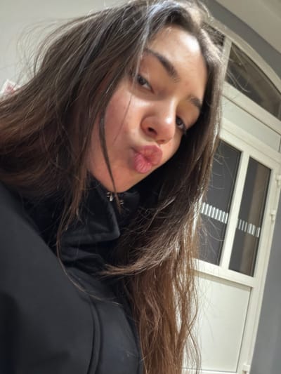 A young woman poses for a selfie with puckered lips. She has long, dark hair and is wearing a black jacket. The background shows a white window with horizontal blinds. The image has a slight tilt and is taken at a close-up angle. The overall tone is casual and playful.