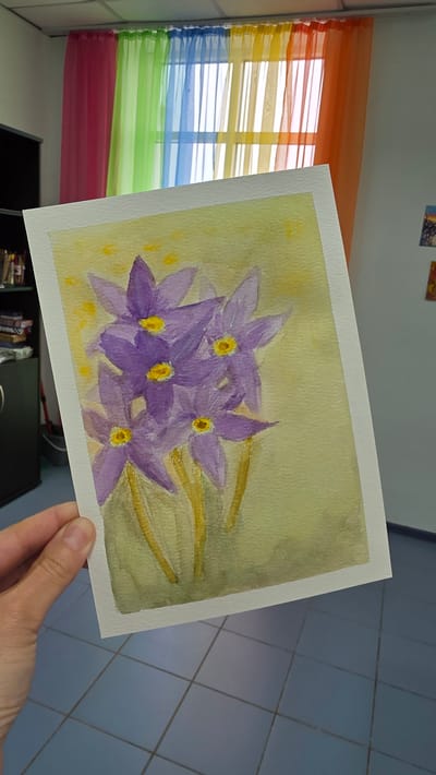 A hand holds a watercolor painting of purple flowers with yellow centers, against a background with a window covered in a rainbow-colored curtain. The painting is on white paper with a visible border. The background shows a tiled floor, a black cabinet, and a corner of a room with a window. The image does not provide information to identify the city of Lensk, Russia.