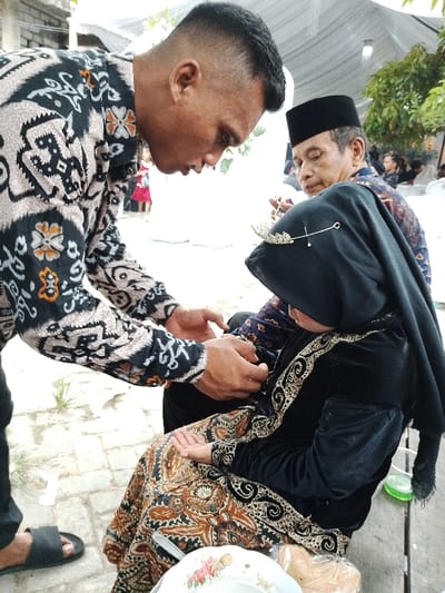 An outdoor scene in Juwana, Indonesia, shows three individuals participating in what appears to be a ceremony or event. In the foreground, a man with short, dark hair, wearing a cream, black, and orange patterned batik long-sleeved shirt and black sandals, leans down. He is using both hands to adjust a detail on the left arm or wrist of a young girl seated in front of him. The girl wears a black headscarf adorned with a gold leaf-shaped pin, a black velvet dress with gold embroidery, and a brown and gold patterned sarong-like garment. She is looking downwards. Behind and to the right of the girl, an older man wearing a black peci (cap) and a blue and brown patterned batik shirt observes the interaction, his right hand resting near his chin.
In the immediate foreground, a white plate with a floral design rests on a wooden surface, holding an item wrapped in clear plastic. A clear cup containing a green liquid is visible nearby. The background features a white canopy or tent structure overhead, green foliage, and blurred figures of other attendees, suggesting a gathering. The ground is a light-colored surface, possibly paved, with sparse weeds visible.