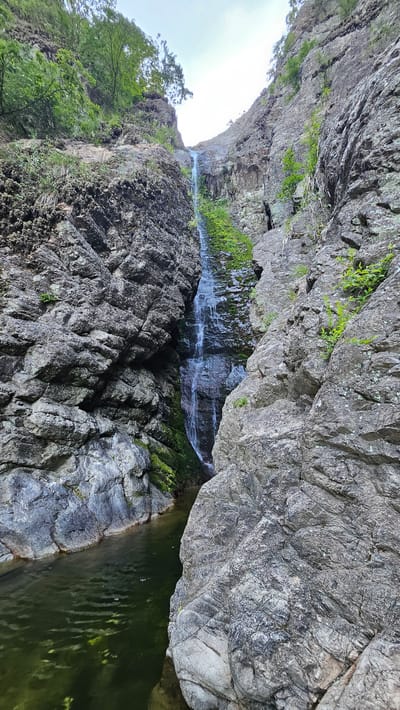 This is a vertical, eye-level, full shot of a narrow waterfall cascading down a rocky gorge. The water flows down a steep, moss-covered rock face, creating a thin stream. At the bottom of the waterfall, a dark green pool of water is visible, surrounded by large, gray rocks. The gorge walls are steep and rough, with sparse vegetation growing from cracks and ledges. The sky is visible at the top, with a slightly overcast appearance. The scene is natural and appears to be in a remote, undeveloped area, possibly a hiking trail or natural park. The image does not contain any people or identifiable man-made structures, aside from the natural formations and water flow.