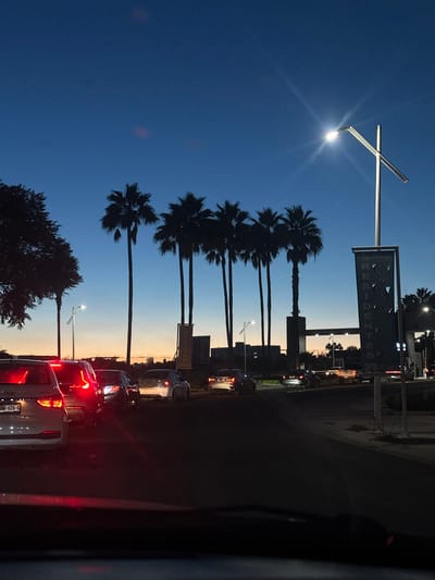 The image depicts a dusk scene in La Purísima, Mexico, showing a multi-lane roadway with active vehicle traffic. The sky transitions from a dark blue overhead to an orange and yellow gradient at the horizon. Silhouetted against the sky are at least seven tall palm trees lining the roadside or a central median, alongside other dark, leafy foliage on the far left.
In the foreground, the image is captured from inside a vehicle, with the dashboard top partially visible. A line of vehicles is stopped or moving slowly, indicated by illuminated red taillights. A white SUV, potentially a Hyundai Tucson with license plate "VMN-626-G," is prominent on the left. Other cars, including a silver sedan and darker vehicles, are visible further down the road.
On the right side of the road, a modern, illuminated street light with a distinctive cross-shaped pole structure casts a bright starburst effect. Beneath this light, a tall, vertical banner partially reads "RESIDENTES." Buildings and distant structures are faintly discernible on the horizon. Smaller, less bright streetlights are also visible in the distance on both sides of the road.
