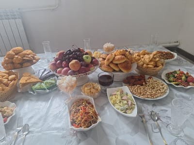 A table laden with a variety of food items is set in a room with a white wall and a radiator visible on the left. The food includes an assortment of pastries, fruits like apples and grapes, nuts, several salads, pasta dishes, and slices of cake. There are also glasses and cutlery on the table, suggesting a meal is being served or has been served. The setting appears to be indoors, possibly a dining area. There are no people visible in the image. The location is described as Новостройка, Kyrgyzstan.