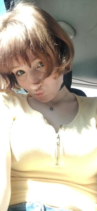 The image displays a head and torso shot of an individual, likely female, situated within what appears to be a vehicle interior. The person has short, light brown hair with bangs, fair skin, and light-colored eyes. A circular metal piercing is present in the nasal septum. A black choker necklace and a silver chain necklace with a small, dark pendant are worn around the neck. The individual is wearing a light yellow, ribbed top with a short zipper closure at the upper chest, and light blue denim material is visible at the bottom of the frame. The lips are pursed in a "duck face" expression, and the gaze is directed towards the camera. The background consists of an out-of-focus light-colored car headliner to the upper right and dark elements suggesting a seat or window frame to the left and behind. Bright light illuminates the subject from the left side, causing some overexposure. There is no discernible visual information within the image to specify a location in the city of Bistrice, Montenegro.