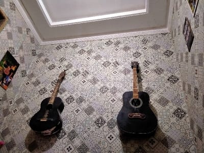 The image shows the interior of a room in a house located at coordinates -6.928, 107.535 in the city of Lagadar, Indonesia. Two acoustic guitars, both black in color, are mounted on the wall, which is covered in patterned wallpaper. The guitars are positioned side-by-side, with the top one slightly higher than the bottom one. The room is dimly lit. There are two picture frames visible on the wall. The room is not occupied, and no one is interacting with the guitars.