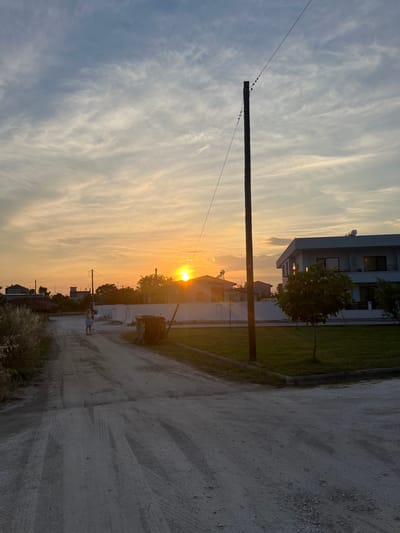 This media file depicts a rural sunset scene in Chaidefto, Greece. The image is oriented vertically. The sky is filled with wispy, elongated clouds, transitioning from a pale blue at the top to a vibrant orange and yellow as it approaches the horizon, indicating the sun is setting. The sun itself is visible as a bright, round orb just above the horizon, casting a warm glow.

In the foreground, a dirt road runs diagonally through the frame. On the left side of the road, a well-maintained lawn is visible, bordering a modern, multi-story house. A small, leafy tree stands in the yard. Further down the road, more houses and trees are silhouetted against the sunset. A wooden utility pole with wires stretches across the sky from left to right, with several small birds perched on the wires near the pole. A lone figure, appearing to be a young woman or girl in a light-colored dress, stands on the road, looking towards the sunset. In the distance, a large, dark dumpster is placed near a building.

The overall mood of the image is peaceful and serene, capturing the beauty of a rural sunset. The time of day is clearly dusk. The weather appears clear with light cloud cover. The presence of houses and a road suggests a residential area or a small village. No visible text is present in the image.