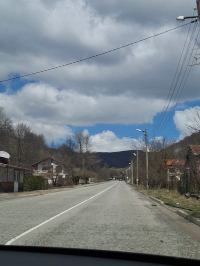 The image depicts a road in Gintsi, Bulgaria. It's a straight, paved road with a white dividing line. Utility poles with wires run parallel to the road. Buildings and trees line the roadside. A mountain is visible in the distance. The sky is partially cloudy.