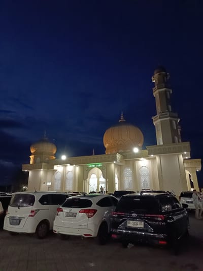This media file shows a mosque illuminated at dusk. The mosque features a large dome and a tall minaret, characteristic of Islamic architecture. Several cars are parked in the foreground, suggesting this is a public space. The sky is a deep blue, indicating the transition from day to night. The lighting on the mosque creates a warm glow, highlighting its intricate details and decorative elements. The text visible on the cars includes license plates and the words "BAITUL NUDA," which likely identifies the mosque. People can be seen near the entrance of the mosque, dressed in casual attire. The scene evokes a sense of peace and community, as people gather at a place of worship during the evening.