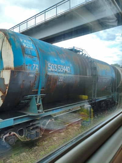 This image captures a section of a blue, weathered tank car on a railway track, viewed through a window. The tank car is large and cylindrical, showing significant rust and peeling paint, indicative of its age and use. White numerals "72" and "503 53440" are visible on its side, along with Cyrillic text that appears to be "РЖД" (RZD), likely indicating Russian Railways. Small, square metal plates are also visible on the surface. The tank car is mounted on a blue chassis with visible wheels and suspension system.

The scene is set outdoors, with railway tracks and gravel visible beneath the train car. Green grass and some vegetation grow alongside the tracks. Above the tank car, a concrete bridge with a metal railing stretches across the frame, suggesting an overpass. The sky is overcast with clouds, hinting at a cloudy day. The lighting is somewhat diffused, with some glare from the window through which the photo was taken. Reflections of yellow and green can be seen on the tank car's surface, possibly from surrounding objects or the environment.

There are no people clearly visible in the frame. The overall mood is somewhat industrial and utilitarian, with the focus on the aged train car. The location context is Buzuluk, Russia, though this is not directly evident from the visual elements alone.