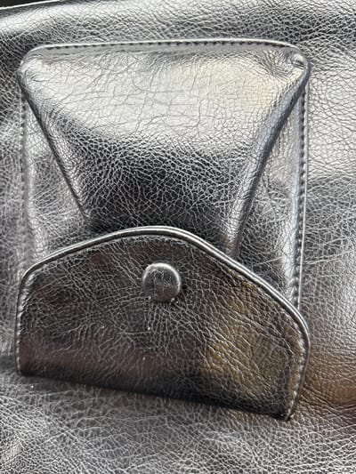 The image is a close-up, high-angle, full shot of a metallic black leather purse. The purse features two pockets: a larger, triangular flap pocket on the upper portion and a smaller, curved flap pocket with a metal button closure below it. The leather has a distinct crinkled texture, and the lighting highlights the sheen of the material, creating strong reflections and shadows. The background is also the same metallic black leather, showing its texture throughout. There are no people or any indication of a city environment present in the image. The location is not specified within the image.
