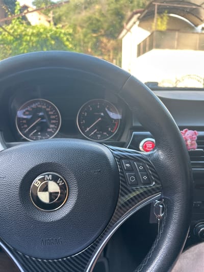 This media file shows the interior of a car, specifically the steering wheel and dashboard. A BMW logo is visible on the steering wheel, along with buttons for various functions. The start/stop engine button is a prominent red circular button. The dashboard includes a speedometer and a tachometer, with visible text indicating speed in kilometers per hour and revolutions per minute. There's also an "AIRBAG" label on the steering wheel. A small pink pig figurine is attached to the car's air vent, adding a whimsical touch. The scene appears to be set during the daytime, as indicated by the natural light filtering into the car. The background visible through the windshield suggests an outdoor setting with trees and some structures. The context provided indicates the location is Djerman, Bulgaria. No people are visible in the frame.