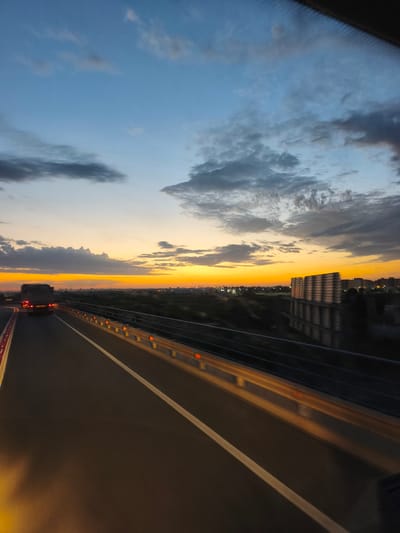 The image depicts a highway at dusk, looking towards the horizon. The sky is a gradient of blues and oranges, with scattered clouds. A truck with its brake lights illuminated is visible on the highway, traveling away from the viewer. The highway has a white dividing line and a safety barrier with orange reflectors. In the distance, a cityscape is discernible with buildings and lights. There is also a large, rectangular structure with a patterned facade to the right of the highway. The scene suggests a moment of travel during twilight, likely on a road leading into or out of a populated area. The specific location is identified as the city of Alcàsser, Spain.