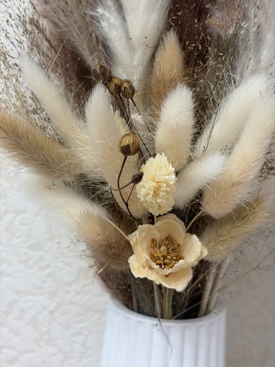 The image displays a close-up shot of a decorative arrangement primarily composed of dried flowers and grasses, presented in neutral, earthy tones. The arrangement is dominated by fluffy, elongated seed heads, likely Lagurus ovatus (bunny tail grass), in varying shades of cream, off-white, and light beige. These are interspersed with thinner, wispy strands of dried grasses in brownish and grayish hues, adding texture and depth to the bouquet.
At the center-right of the arrangement, there are two distinct dried flowers. The larger one is an open bloom, delicate and light cream in color, with visible stamens or a central cluster of petals in a slightly darker beige. To its left, a smaller, spherical flower with a tightly clustered, intricate petal structure is visible, in a pale yellow or off-white shade. Below and to the left of these flowers, a cluster of small, round, dried seed pods or berries, dark brown in color, are attached to thin, wiry stems.
The overall color palette is very subdued, consisting mainly of whites, creams, beiges, light browns, and grays, contributing to a rustic and natural aesthetic. The textures are diverse, ranging from the soft, feathery quality of the fluffy grasses to the delicate dryness of the flowers and the smooth, hard appearance of the seed pods.
On the far right side of the image, a portion of a white, vertically ribbed object is visible, suggesting it might be a vase or a decorative container holding the arrangement. The background is softly lit and appears to be a light-colored, possibly textured wall or surface, further emphasizing the foreground elements. The lighting is soft and even, highlighting the intricate details and textures of the dried botanical elements.
