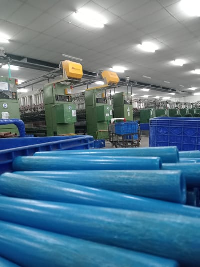 The image captures an interior view of an industrial manufacturing facility, identified as being located in Nglundo, Indonesia. The foreground features a close-up, slightly out-of-focus stack of numerous cylindrical blue objects, consistent with empty bobbins or yarn cones. Partially visible blue plastic crates are also present in the immediate foreground.

In the midground and background, a large, well-lit factory floor is visible, populated by multiple rows of heavy industrial machinery. These machines are predominantly green in color and appear to be textile or yarn processing equipment, featuring numerous spindles, some holding white spools of yarn. Several machines display yellow overhead units with the "WAMSLER" brand visible. Individual machines are marked with numbers such as '12', '13', '14', and '15'. Blue plastic crates are interspersed throughout the machine rows, with one crate positioned on a wheeled cart. A blue pipe or conduit is visible on the left side, extending from one of the green machines.

The facility's ceiling is white, constructed from square acoustic tiles, and is extensively illuminated by multiple recessed rectangular fluorescent light fixtures. Overhead ductwork or conveyor systems run above the machinery. No individuals are visible within the scene.