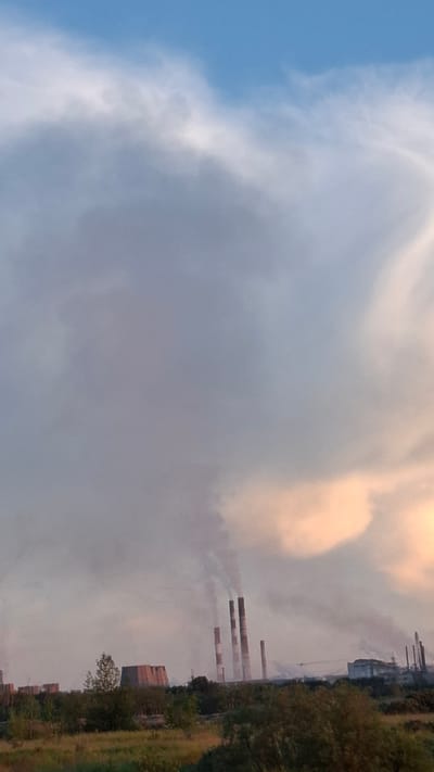 The image depicts an industrial landscape under a sky filled with clouds. The primary subjects are several tall smokestacks emitting plumes of smoke, indicating industrial activity. These smokestacks are positioned along a treeline on the right side of the frame. In the background, industrial buildings and structures are visible, some appearing to be factories or power plants. The foreground on the right shows a grassy area with some trees. The sky is a mix of blue, grey, and a warm, peachy hue, suggesting either sunrise or sunset. The lighting and cloud cover create a diffused atmosphere. The overall scene conveys a sense of industrial operations within a natural or semi-natural setting. The orientation of the image is landscape, with the horizon running vertically along the right edge. The location is identified as Achinsk, Russia.