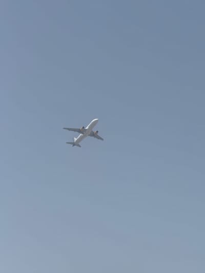 An airplane is visible in a clear, pale blue sky. The airplane is white with yellow engine nacelles and a gray underside with some markings. It is captured from below, angled upwards as if ascending. The image does not depict any urban elements, indicating that the aircraft is likely in flight above an area such as Guadalmar-San Julián, Spain, rather than within the city itself. No people or ground-based objects are discernible.