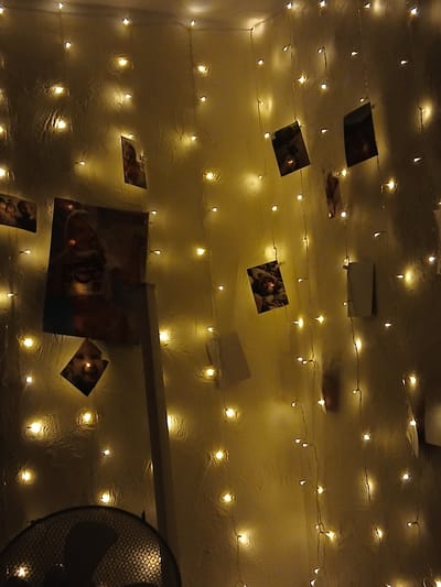 The image depicts a dimly lit room, likely a bedroom or living area, decorated with strings of warm, yellow fairy lights. The lights are draped vertically and horizontally across a textured, light-colored wall, creating a cozy and atmospheric ambiance. Several photographs and possibly other small prints are attached to the wall, interspersed among the lights. One prominent photograph, a poster-sized image, features a person with blonde hair, wearing a white top with a cherry design. Another smaller, diamond-shaped photograph shows a baby. Other visible items include a dark, metallic fan partially visible at the bottom left of the frame and a white vertical support structure on the right side of the poster. The overall scene is one of personal decoration and a relaxed, intimate setting. The mention of Rukla, Lithuania, suggests a potential location context for this scene, though the image itself does not contain any direct visual cues to confirm this specific location.