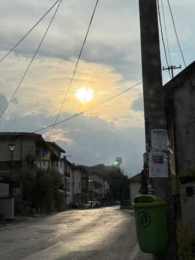 The image depicts a street in Konjic, Bosnia and Herzegovina, with buildings lining both sides of the road. The sun is setting, casting a warm glow on the wet asphalt and clouds. Overhead, electrical wires crisscross the sky. A green trash can with a recycling symbol is visible on the right side of the street, attached to a utility pole adorned with posters. A flag, likely the flag of Bosnia and Herzegovina, is displayed on one of the buildings on the left. Several cars are parked or driving on the street. The scene appears to be a residential or commercial area.