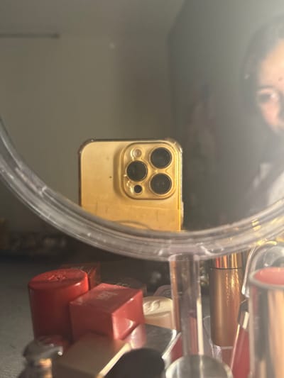 A person’s reflection appears in a round mirror. Within the mirror’s reflection, a gold iPhone with three rear cameras is prominent. The mirror is surrounded by various items, including several lipsticks, some in red packaging, creating a cluttered border around the reflective surface.