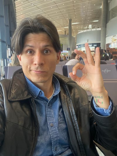 This image depicts a male individual taking a selfie within Tocumen International Airport, located in Tocumen, Panama.

The subject, a male with dark hair parted in the middle, dark eyes, and light-to-medium skin tone, occupies the foreground. He is wearing a black leather jacket over a blue denim button-up shirt, with a light-colored undershirt and a thin silver-colored chain necklace visible. His expression features wide eyes and a slight smirk. His right hand is raised to chest level, forming an "OK" gesture with the thumb and index finger creating a circle, and the other three fingers extended. On the inside of his right wrist, a tattoo is visible, displaying the word "Ditch" (oriented to be read when the arm is rotated) followed by green and red candlestick chart symbols.

The background reveals a spacious, well-lit airport terminal. High ceilings with a grid pattern and numerous recessed lights are visible. Large windows line the left side, admitting natural light. Rows of grey airport seating are present, some occupied by other indistinct individuals. Crucially, a sign clearly displaying "Tocumen" is visible on the back of one of these seats, confirming the location as Tocumen International Airport. Further in the background, commercial signage, including a partial "shop" sign and a yellow sign marked with the number "1," is present, along with architectural columns and other distant figures.