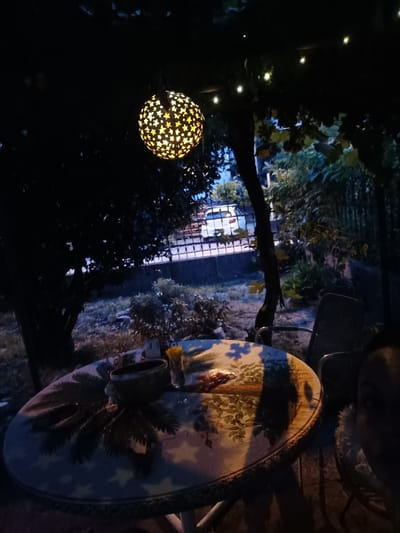 This is a dimly lit, outdoor scene, likely a backyard or patio at dusk or night. In the foreground, a round table is covered with a patterned cloth. On the table are a dark bowl, a small vase with yellow flowers, and what appears to be some food items. To the right, partially visible, is a person. In the background, a white car is parked behind a decorative metal fence. The area is surrounded by trees and foliage, with string lights visible overhead, including a decorative spherical lantern with star cutouts hanging from a branch. The environment suggests a residential area in Serravalle, Switzerland.