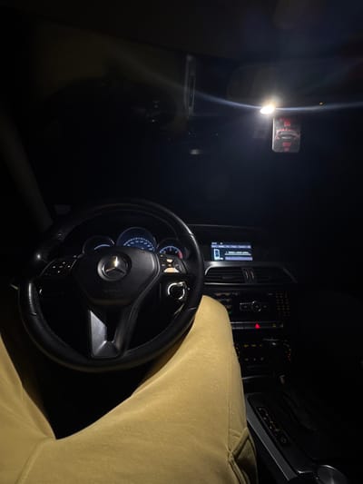 An interior shot of a vehicle, taken from the driver's perspective at night or in a dark environment, located in Vranentsi, Bulgaria. The driver's legs, clad in mustard yellow or khaki-colored sweatpants, are visible in the foreground. A black, multi-functional Mercedes-Benz steering wheel is centered. The illuminated instrument cluster shows low or zero speed and RPM readings. The center console's infotainment screen displays "19:04," "BULGARIA," and "TRAP GOD KRAB LEVNA" along with a phone icon, indicating media playback. Below the screen are air vents, climate controls, and other buttons, including a red hazard light button. An automatic transmission gear selector is visible to the right of the driver's leg. Overhead, a bright interior light is illuminated, casting light on a dashboard camera mounted near the rearview mirror and a red air freshener with "G6" or "G0" visible, hanging from the mirror.