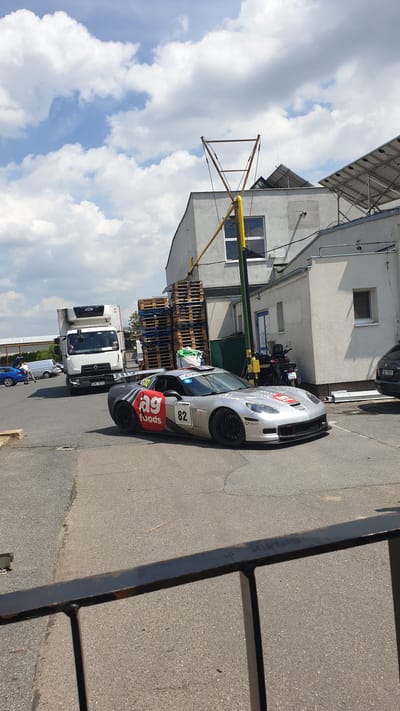 The image depicts a silver Chevrolet Corvette sports car with racing decals, including "ag foods" and the number "82", parked on an asphalt surface in front of a light-colored industrial building. A white truck is visible to the left of the sports car. Several stacks of wooden pallets are piled near the building. The sky is partly cloudy. The scene appears to be in an outdoor industrial or commercial area, consistent with the provided location of Vestec, Czechia.