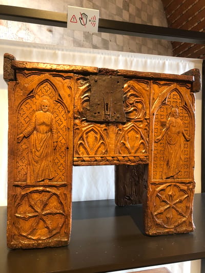 A detailed, ornate wooden carving is displayed in a museum setting. The carving is light brown and appears to be quite old. It features two similar rectangular sections positioned horizontally, with a connecting vertical section. Each horizontal section is decorated with a relief carving of a figure, possibly religious, surrounded by ornate patterns. The top and bottom horizontal sections feature a relief carving with a similar pattern. There is a metal latch or lock attached to the center vertical section, indicating the piece may have been part of a larger structure, such as a chest or door. The object is exhibited in a glass display case, with a white backdrop. To the left of the display, there is a warning sign with an image of a hand with a line through it, suggesting "Do Not Touch". The floor near the object has a checkered pattern. The location is Château de Chillon, Veytaux, Switzerland.