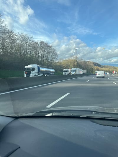 The image depicts a multi-lane highway in Beausemblant, France, observed from inside a vehicle, with the dashboard and lower windshield visible in the immediate foreground. The asphalt highway features white lane markings, indicating at least two lanes of travel in the depicted direction. To the left, separated by a low concrete barrier, a grass verge or shoulder hosts several stationary heavy goods vehicles. The most prominent is a white MAN truck tractor coupled to a metallic silver tanker trailer, which bears partially visible blue text including "H2 MOBILISER ENSEMBLE." Behind this tanker truck, at least two additional white tractor-trailer trucks are also stopped. In the right-most lane of the active highway, a white panel van is visible traveling further ahead, with other vehicle traffic receding into the distance. The environment beyond the highway shoulder includes a line of deciduous trees, mostly bare, suggesting late autumn or early spring. A large white wind turbine is prominently positioned on elevated terrain behind these trees to the left. Distant hills are discernible on the horizon. The sky is partly cloudy, displaying patches of blue interspersed with white clouds. A yellow, elongated object, consistent with a kite, is visible in the upper right portion of the sky. No human figures are clearly identifiable.
