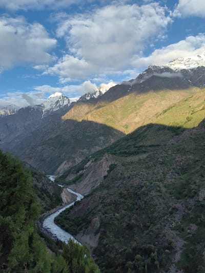 This media file is a photograph capturing a majestic mountain landscape with a winding river. The scene is set in Stingri, India. The primary subjects are the dramatic, rugged mountains, some of which are snow-capped, indicating a high altitude. A vibrant blue sky with scattered white clouds dominates the upper portion of the image, suggesting fair weather.
A significant detail is the interplay of light and shadow across the mountain slopes. Sunlight illuminates some areas with a warm, golden hue, highlighting the green vegetation and rocky terrain, while deep shadows carve out the contours of the mountains, adding depth and drama to the scene. The time of day appears to be late afternoon or early evening, as evidenced by the angled sunlight casting long shadows.
A prominent feature of the landscape is a turquoise river that snakes through a deep valley. The river appears to be fast-flowing, with white water indicating its turbulence. The valley walls are steep and covered in a mix of rocks and sparse vegetation, with some trees visible, particularly in the foreground. The overall mood of the image is one of awe-inspiring natural beauty and tranquility, conveying a sense of remoteness and the grandeur of the Himalayas. There is no visible text in the image.