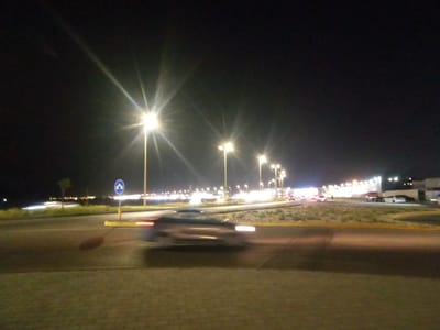 The image presents a nighttime scene on a road in Al Khobar, Saudi Arabia. The dark sky is dominated by numerous bright, starburst-effect streetlights that cast a strong yellow-orange illumination over the area.
In the foreground, a paved surface, likely a sidewalk or shoulder, is visible. The main subject of the mid-ground is a road where a vehicle is in motion, depicted with significant motion blur, appearing as a dark, streaking shape with discernible red taillights. A blue circular road sign with a white upward-pointing chevron arrow stands beside the road, indicating a traffic-related instruction.
The road features a median strip with low, sparse vegetation. In the background, a continuous line of lights suggests distant traffic or buildings, contributing to an urban or suburban nighttime environment. Other vehicles, some stationary and some in motion with blurred lights, are visible further down the road. There are no discernible people or visible text in the frame. The overall impression is one of active transit under strong artificial lighting at night.