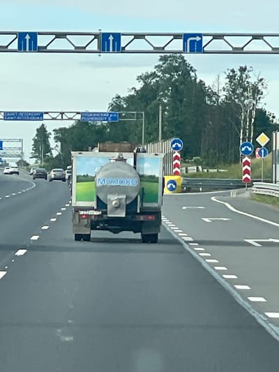 The image captures a view of a highway in the vicinity of Vyra, Russia. In the foreground, a white milk tanker truck is driving on the asphalt road. The truck has the word "МОЛОКО" (MILK) written in blue Cyrillic letters on its cylindrical tank, which is decorated with a scenic image of a green field and a blue sky. Several other vehicles, including cars, are visible on the road ahead of the truck. Overhead, a metal gantry displays various road signs, including directional arrows indicating straight ahead and a right turn. The surrounding environment consists of trees and vegetation. The sky is overcast.