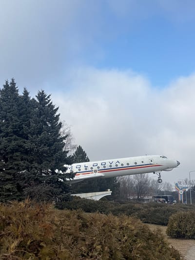 An Ilyushin Il-18 passenger aircraft, predominantly white with "MOLDOVA" in blue capital letters and a red, white, and blue stripe featuring a national emblem on its fuselage, is displayed as a monument. The aircraft is mounted on a light-colored concrete support structure, positioned at an upward angle suggestive of flight, with its nose landing gear extended. This scene is located at the entrance to Chisinau, specifically near Bubuieci, Moldova.

In the foreground, dense, low-lying shrubs with brownish-green foliage cover the ground. To the left, a cluster of tall, dark green coniferous trees partially obscures the aircraft's wing and tail section. Beyond these, bare deciduous trees are visible in the mid-ground. To the right of the aircraft's nose, a large, multi-colored welcome sign, featuring blue, white, and yellow elements, clearly displays the word "CHISINAU." Behind this sign, a dark-colored bus is visible. The sky above is partially cloudy, presenting a mix of light blue patches and white-grey clouds, indicating daylight conditions. No individuals are visible.