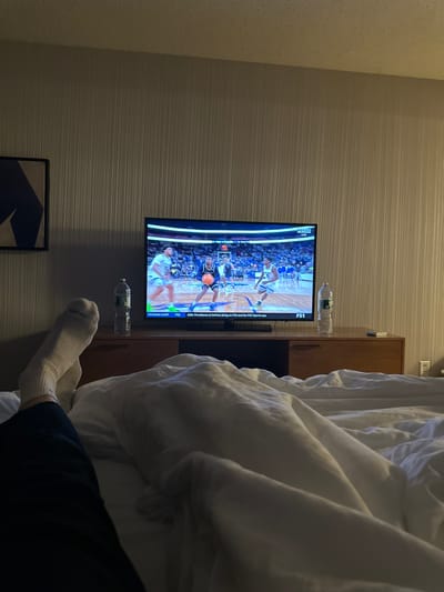 A person lies in bed watching a basketball game on television. The bed is covered in white sheets and a comforter. The person is wearing socks and dark pants. Two water bottles sit on the dresser to the side of the television. The wall has a lined wallpaper and a framed piece of art is visible.