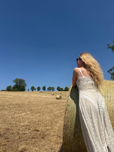 In Tandel, Luxembourg, a blonde woman wearing a white lace dress and sunglasses leans against a large, round hay bale in a sunlit, harvested field. The sky above is a clear, vibrant blue, with a few distant trees visible on a gentle slope in the background. Several other hay bales are scattered across the dry, golden field, suggesting it's late summer or early autumn. The woman is facing away from the camera, looking out towards the horizon, her long, wavy blonde hair blowing in the breeze. The scene evokes a sense of rural tranquility and a bright, warm day.