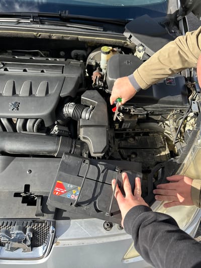 In Fegersheim, France, two individuals are engaged in automotive maintenance on the open engine bay of a silver Peugeot vehicle during daylight. One person's right hand, wearing a khaki jacket, operates a green and red handled screwdriver near a black electrical component housing. Simultaneously, another person's hands, wearing a dark long-sleeved garment, are positioned on a black plastic cover bearing a 'LiB - 70Ah' label and various battery warning symbols, indicating it is a battery component. Visible engine elements include the Peugeot-branded engine cover, air intake system, and fluid reservoirs.