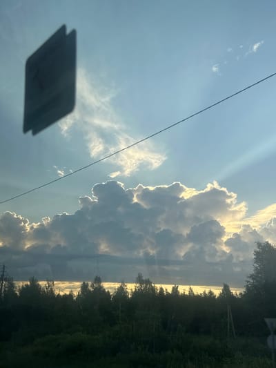 A partially obscured, dark rectangular object, possibly a sign or sticker, is affixed to a glass surface, casting a shadow. Beyond this surface, a wide expanse of the sky dominates the frame, featuring a blend of light blue and white clouds. Dramatic sunbeams pierce through the clouds, illuminating them from behind and creating a radiant halo effect. A thin, dark horizontal line, likely a wire, stretches across the upper portion of the sky. The lower third of the image reveals a dense treeline of varying heights, with a strip of hazy, yellow light visible above the trees. The ground in the foreground is covered in dark vegetation. The scene appears to be captured outdoors, likely from a moving vehicle given the perspective and the presence of the object on the glass. The location is specified as Yarkovo, Russia.
