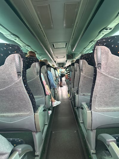 The image shows the interior of a tour bus, likely in Fuengirola, Spain, with passengers seated. The viewpoint is from one of the seats, looking down the aisle of the bus. On the right, a row of grey fabric seats with a black pattern of small orange and white dots is visible. Each seat has grey plastic armrests and supports. The aisle floor is dark grey. On the left, the back of a grey fabric seat with the same black pattern is partially visible. In the foreground on the left, the back of another seat and a glimpse of a person's legs are seen. Further down the aisle, several passengers are visible, some with their backs to the camera and others seen from the side. One passenger in the distance is standing and appears to be talking to someone. The ceiling of the bus is light grey and features air vents and lighting fixtures. A small screen is mounted on the ceiling towards the front of the bus. The overall impression is of a typical bus interior, suggesting travel or a guided tour.