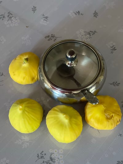 The image depicts a still life arrangement of five yellow figs and a metallic sugar bowl with a glass lid on a grey tablecloth with a subtle floral pattern. The figs are round and plump, with visible stem remnants on some. The sugar bowl is round and shiny, with a spoon resting inside. The tablecloth has a repeating pattern of small white flowers and grey sprigs. The lighting is even, suggesting an indoor setting. The location provided, Tasbuget, Kazakhstan, is not visually evident from the image itself.