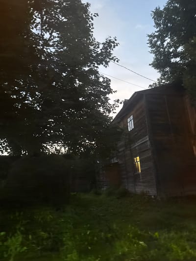 An outdoor, low-angle, wide shot captures a wooden, multi-story building adjacent to a dense foliage of trees. The sky, visible in the upper section, transitions from a light blue to a darker hue, suggesting either early morning or late evening. The building's facade, constructed from horizontal wooden planks, displays two windows. One window on the upper floor is illuminated with a bright white light, while a window on the lower floor emits a warm yellow glow. A power line stretches across the frame, diagonally from the upper left to the upper right. The foreground is dominated by lush green grass and the shadows of the surrounding trees, creating a somewhat dark and moody atmosphere. The overall scene appears to be located in a rural or semi-rural area, possibly within the city of Vereya, Russia, given the architectural style and natural surroundings. There are no people or animals visible in the image.