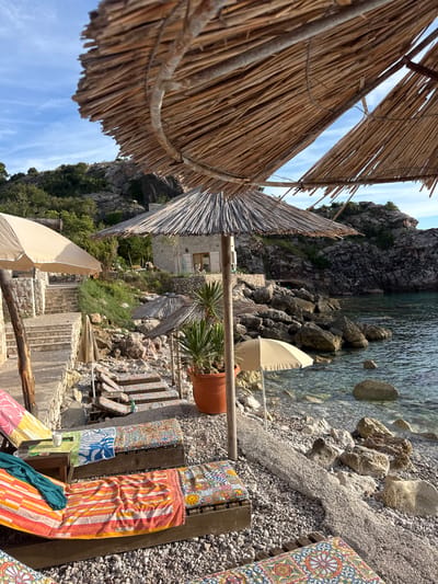 The image depicts a tranquil beach scene in Rijeka Reževići, Montenegro. The foreground features several wooden loungers, each adorned with vibrantly colored towels and patterned cushions, suggesting a comfortable and inviting atmosphere. Behind the loungers, a pebble beach slopes down to the clear, calm water of the Adriatic Sea, which is dotted with large rocks. The mid-ground reveals more beach chairs and a few straw umbrellas providing shade. A stone pathway with steps ascends the hillside, leading towards a small, rustic stone building. Lush green vegetation and rocky outcrops characterize the rugged coastline in the background. The sky is a clear blue with a few wispy clouds, and the sunlight casts long shadows, indicating it is late afternoon or early evening. No people are visible in the frame.