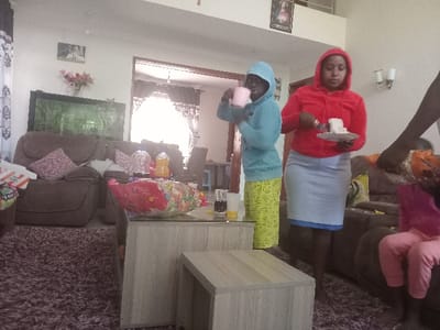 The scene is identified as being in Syokimau, Kenya. The image depicts an indoor domestic setting, specifically a living room, furnished with brown plush sofas and a dark purple shag rug. Two light-colored, multi-tiered coffee tables with glass surfaces are positioned in the foreground. On the larger coffee table, a clear bag of colorful snacks, two plastic bottles containing dark and orange liquids respectively, and a glass with an amber liquid are visible.

Three adults and one child are present. A person on the left wears a light blue hooded sweatshirt and yellow patterned trousers, holding a pink mug to their mouth. To their right, a person in a red hooded sweatshirt and a light blue skirt holds a white plate with a slice of white cake and a fork. On the far right, a partial view of an adult's arm in an orange and brown patterned garment is seen, reaching towards a child seated on a brown sofa. The child is wearing a pink top and pink trousers.

The room's walls are light-colored, adorned with a framed religious image above an archway in the background, another framed picture on the left wall, and two wall sconces on the right. A large, dark-framed aquarium is situated behind the left sofa. Pink artificial flowers in a vase are visible on a side table on the left. Patterned curtains are present on a window on the left and around the background archway. The scene is well-lit, indicating daytime.